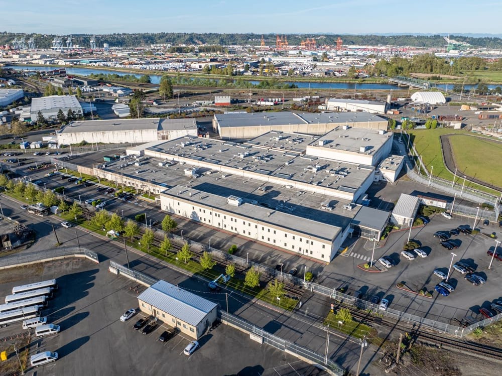 An aerial drone image shows a two-story warehouse like detention center with the tower cranes of the Port of Tacoma in the background.