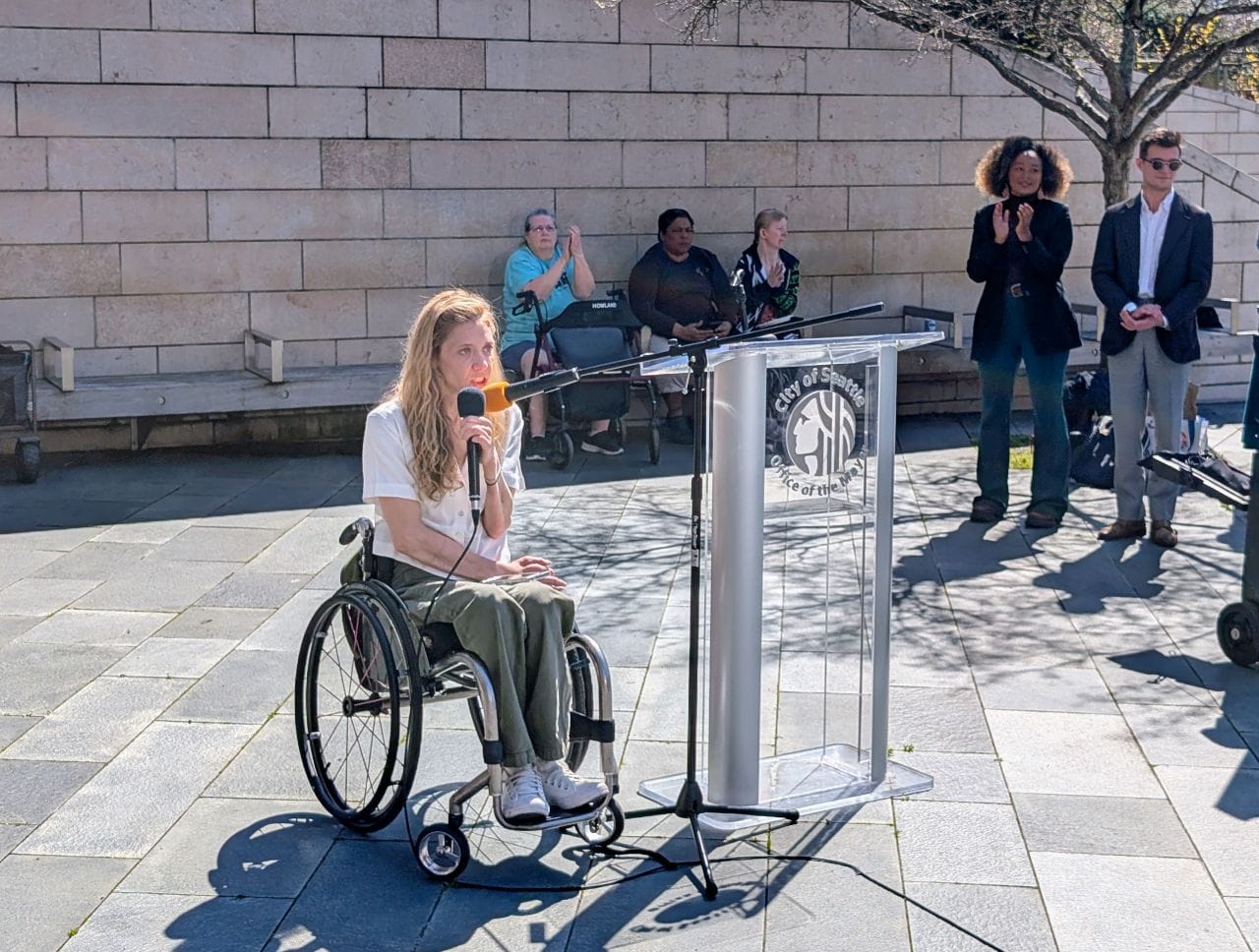 Cecilia is a wheelchair user and holds the mic while housing advocates in the background clap for the point she made.