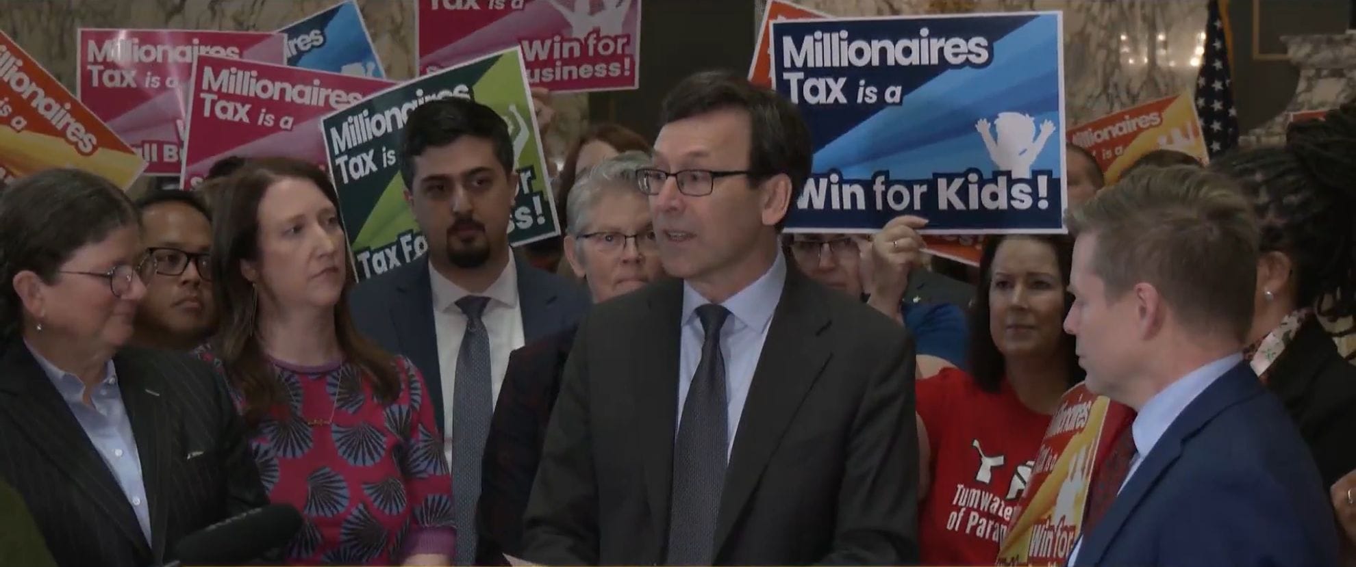 Supporters hold sign saying "Millionaires Tax is a win for kids!" and top lawmakers stand beside Ferguson with Senator Noel Frame to the left and Jamie Pedersen to the right.
