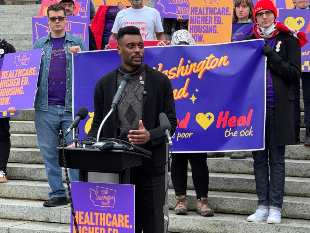 Scott stood at a lectern with supporters behind him up the steps with signs saying "healthcare. higher ed. housing. now."