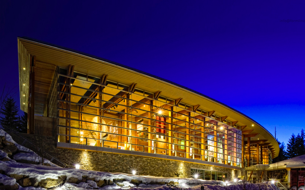 A photo of a curved wooden building lit up against a blue sky. 