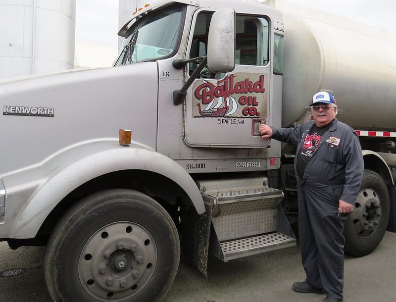 Mustached man standing in front of an oil truck with Ballard Oil Co written on the door