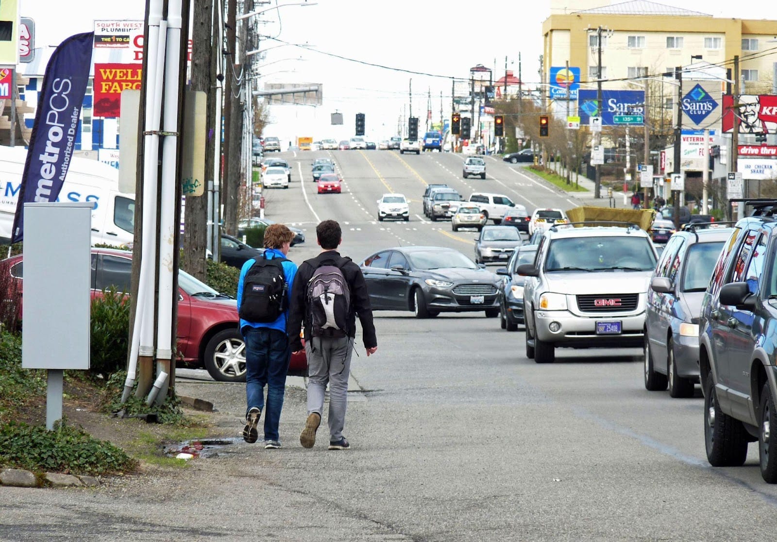 Rows of cars on a block of drive thrus and strip malls as two kids walk in the gutter of the road.