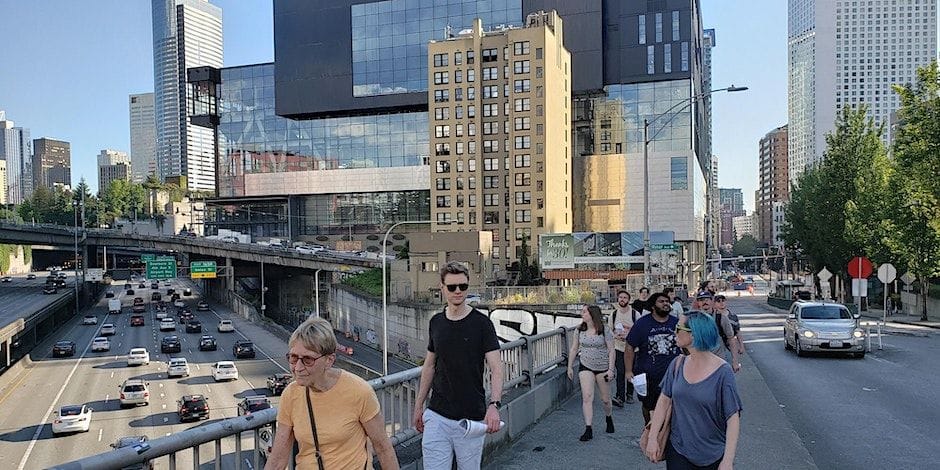 A line of a couple dozen people walk on Pike/Pine corridor over I-5 with many cars in the trench below.