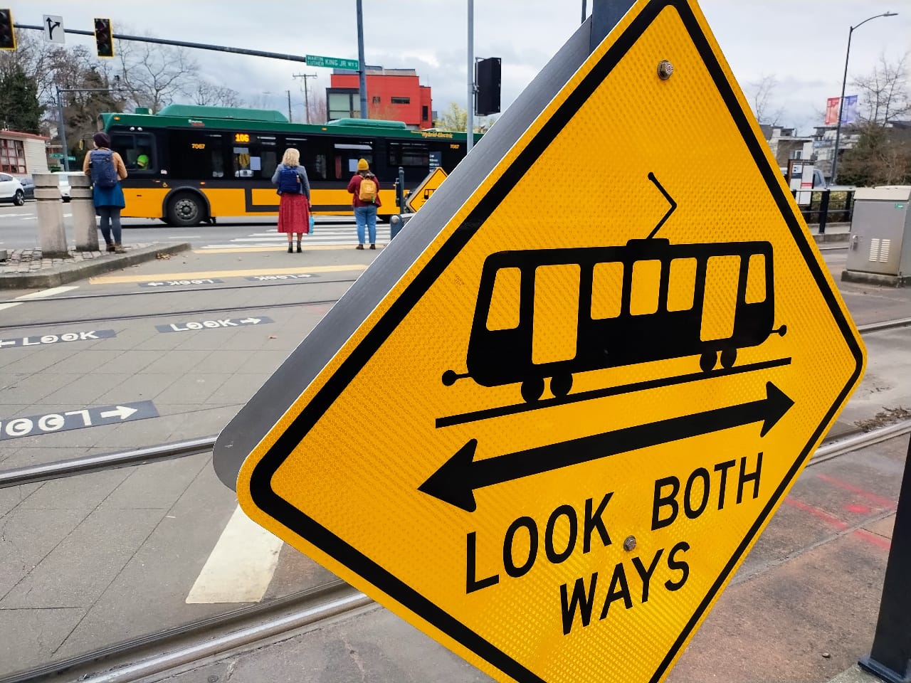 A look both ways yellow sign includes a light rail symbol. Pedestrian wait at the opposite corners of MLK Way.