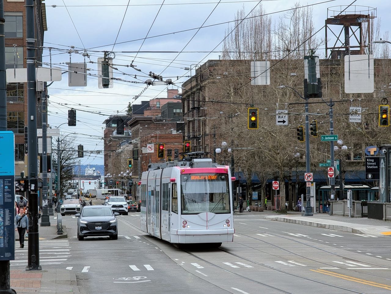 A white streetcar runs down the middle lane on Jackson Street next to King Street Station. Pedestrians walk on the sidewalk and cars run alongside 