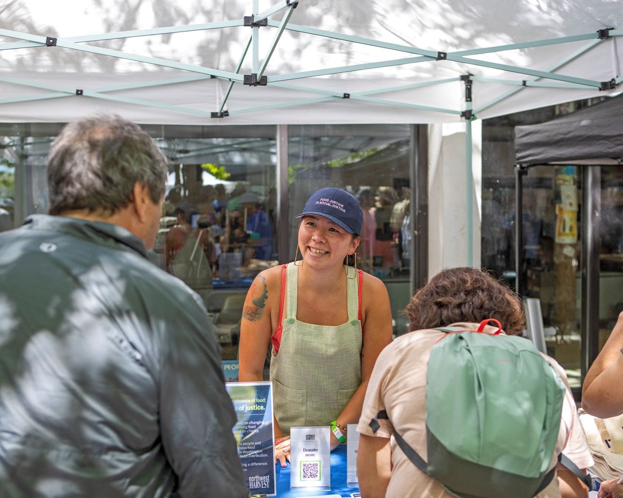 Chunn stands at a table in an apron with Northwest Harvest informational materials strewn on the table chatting with two guests.