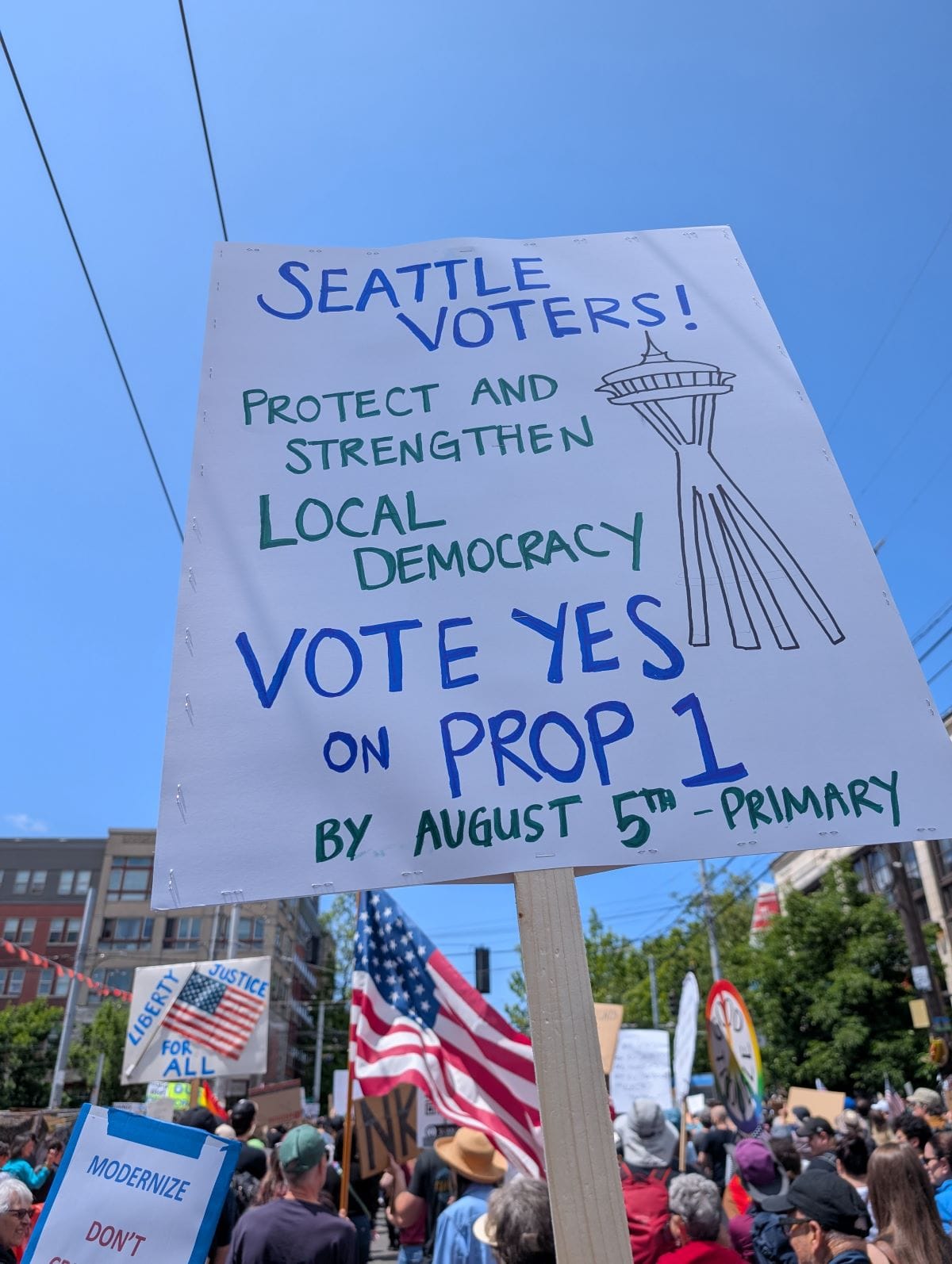 Amidst a crowd of marchers, a sign reads "Seattle voters! Protect and strengthen local democracy. Vote yes on Prop 1 by August 5th Primary."