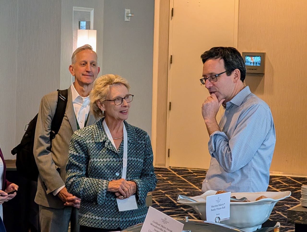 Ferguson grips his chin and he converse with Gregoire. A buffet table has a platter of potato rolls near where they stand.