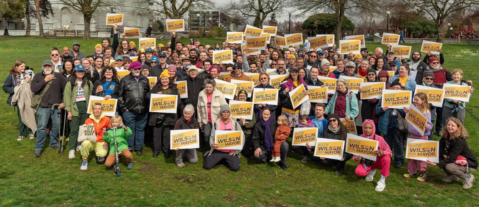 Nearly 100 campaign supporters stand in a grassy park waving yellow Wilson campaign signs.