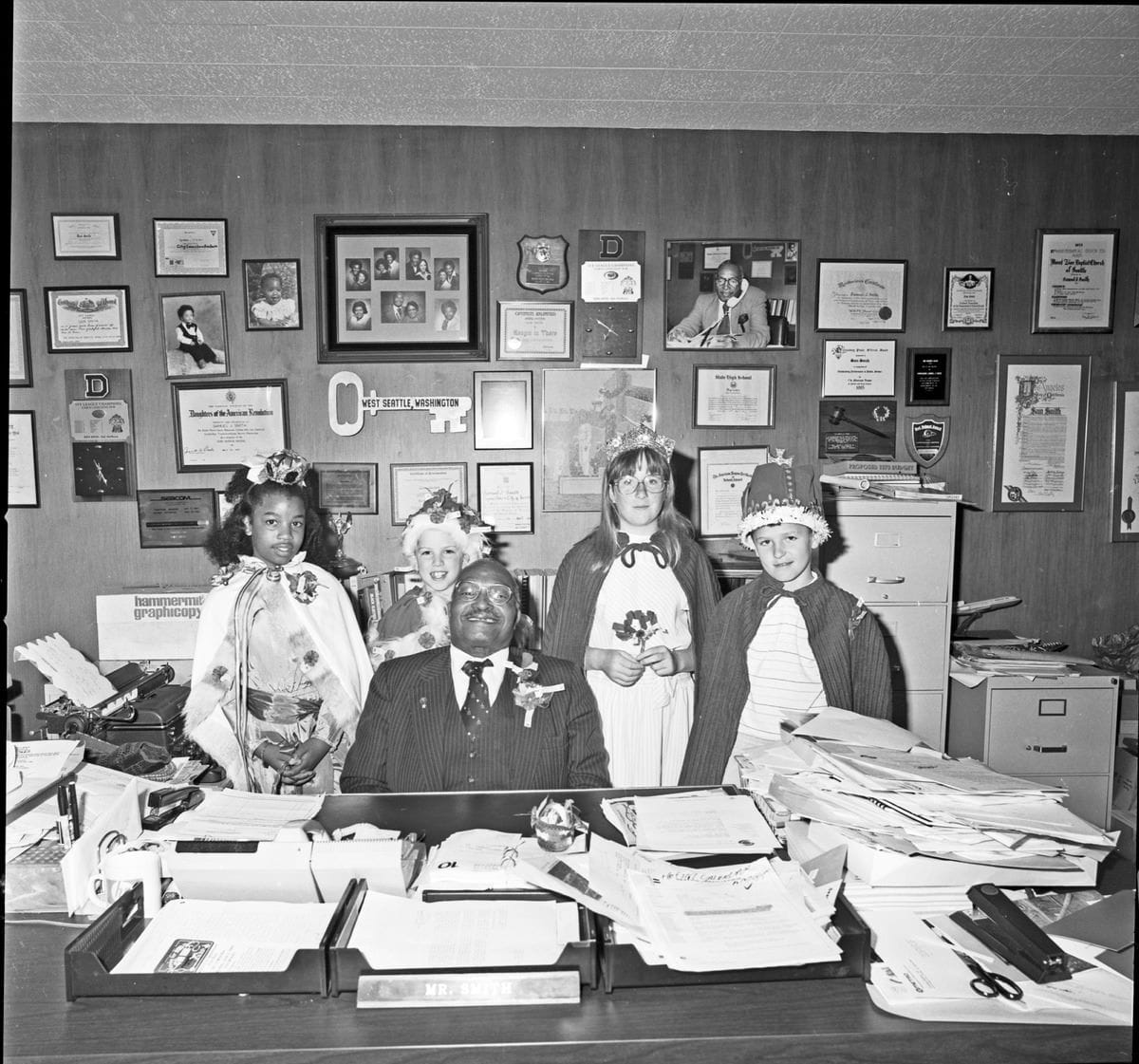 Smith wears a suit and sits at a desk with stacks of papers and the wall behind him is filled with accolades and photographs. 