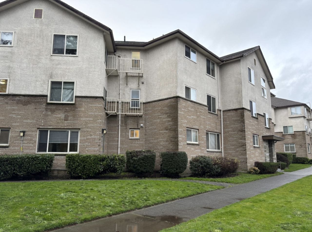 A three-story apartment building with light-colored brick and beige siding and a grass lawn