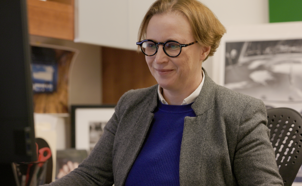 Belman is light-haird and wears glasses and a blazer, sitting at an office chair.