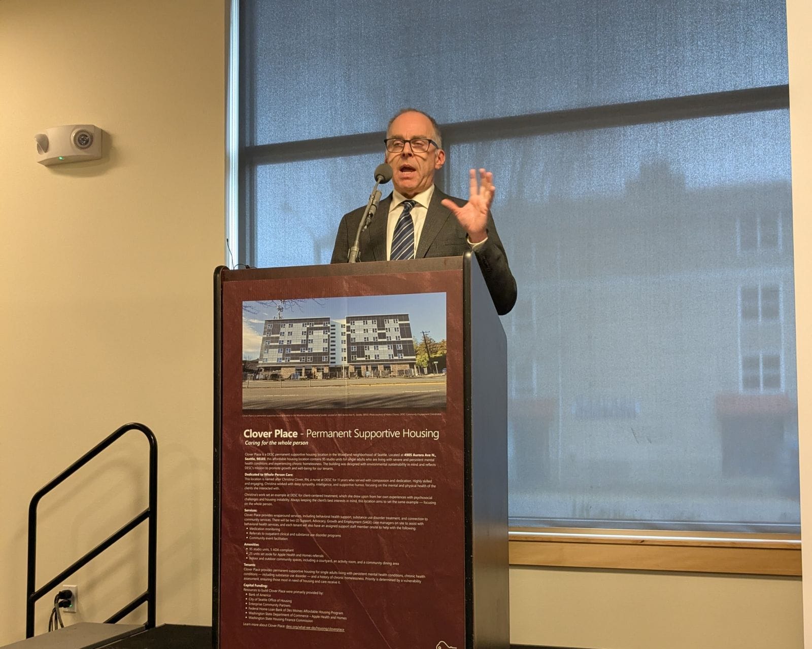 Malone wears a navy-colored suit and stands at a lectern with a picture and description of Clover Place on a  posterboard