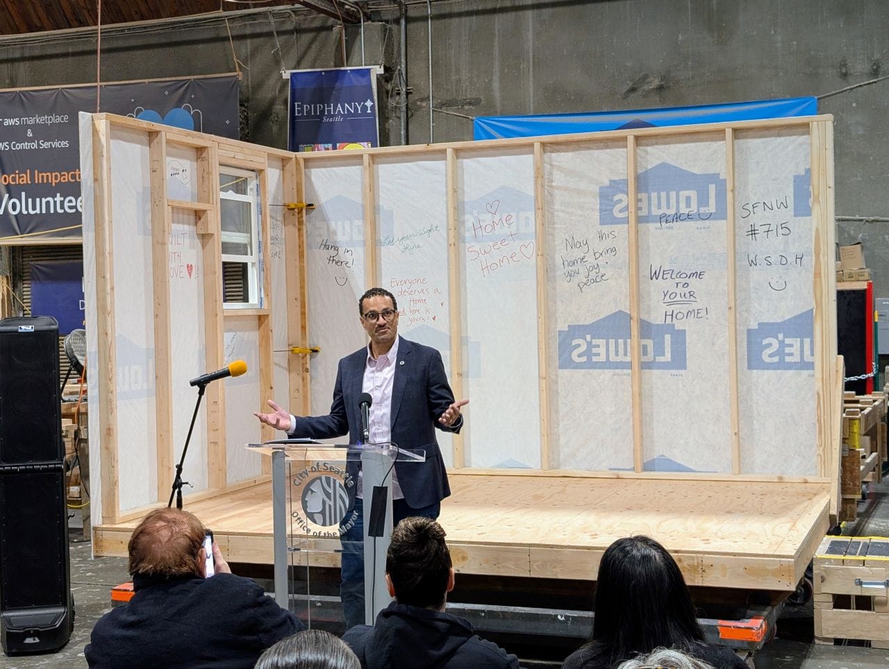 Saka wears a suitcoat and stands in front of a half-completed tiny house in a factory.