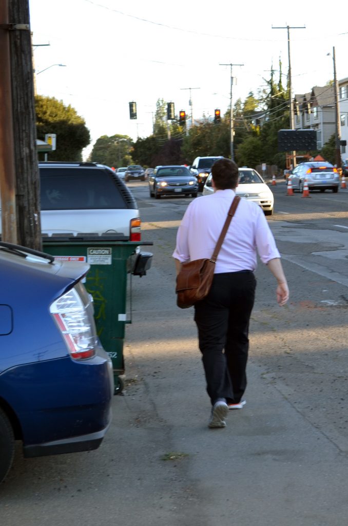 A man walks between parked cars and oncoming traffic on Greenwood Avenue N. (photo by author)