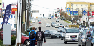 Two teens walk along the edge of Aurora with no sidewalk and streams of cars going by.
