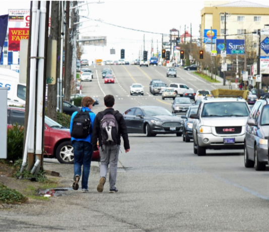Fix Aurora Avenue Now Two teens walk along the edge of Aurora with no sidewalk and streams of cars going by.
