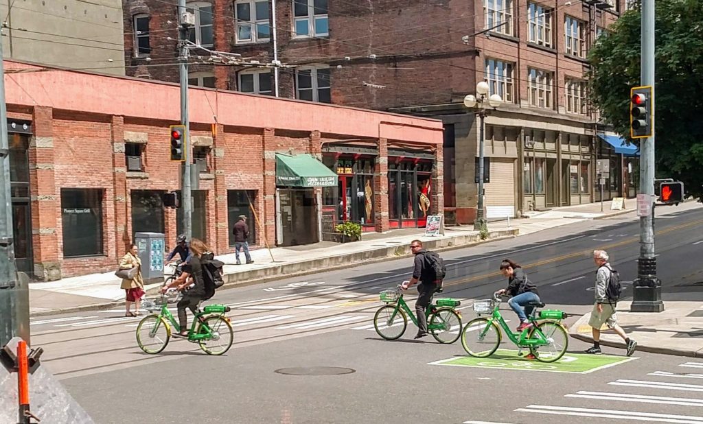 Three people ride through an intersection in Pioneer Square on old-school Lime bikes with battery packs on the back