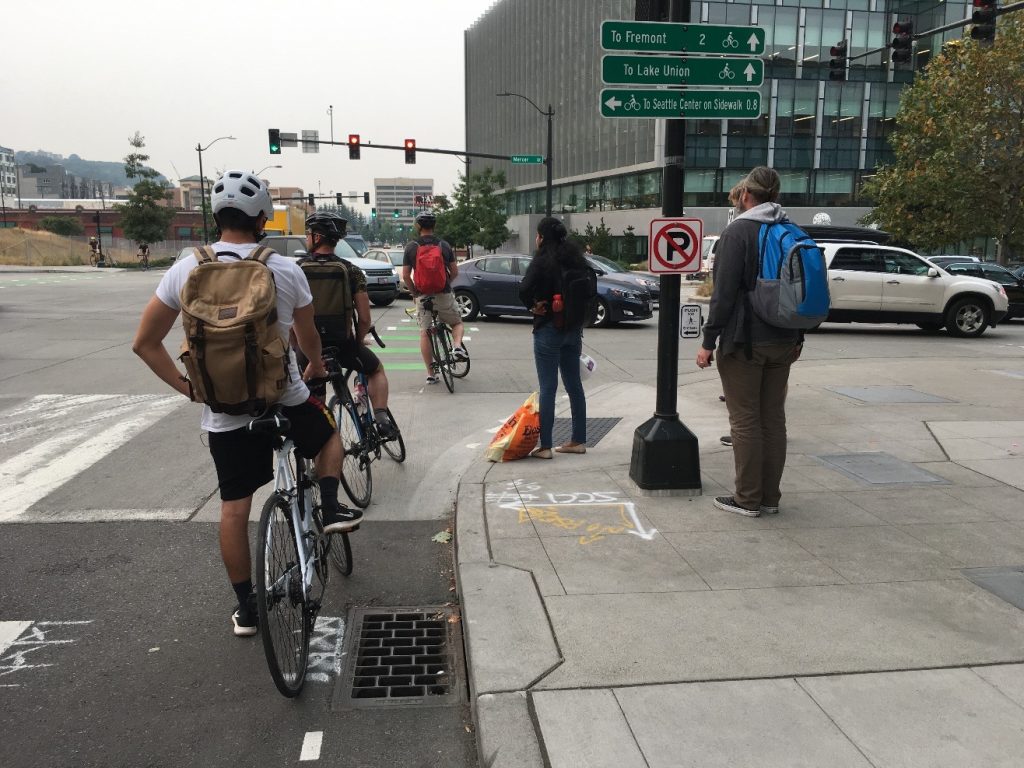 Three people biking and two pedestrians wait for a long line of cars queuing on Mercer Street in South Lake Union.