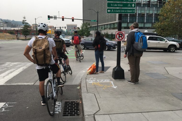 Three people biking and two pedestrians wait for a long line of cars queuing on Mercer Street in South Lake Union.