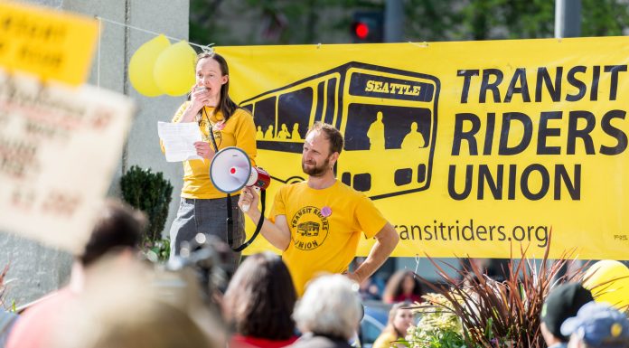 How Mayor-elect Wilson Can Hit the Ground Running, Walking, Rolling, and Biking Wilson holds a bullhorn and speaks at a pre-pandemic rally standing in from or a big yellow Transit Riders Union banner.
