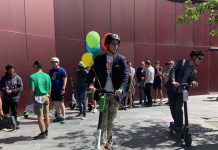 For a Brief Moment, Scooters Took Over Seattle’s City Council Plaza A dozen folks stand around as two people test ride e-scooters. Balloons adorn the city hall plaza and its burgundy paneled wall.