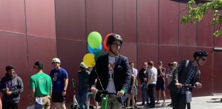 A dozen folks stand around as two people test ride e-scooters. Balloons adorn the city hall plaza and its burgundy paneled wall.