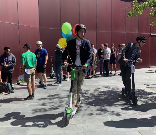 For a Brief Moment, Scooters Took Over Seattle’s City Council Plaza A dozen folks stand around as two people test ride e-scooters. Balloons adorn the city hall plaza and its burgundy paneled wall.