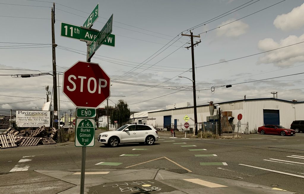 A photo showing a stop sign and a sign indicating the end of the Burke Gilman Trail. The street behind it shows a car turning into the intersection. 