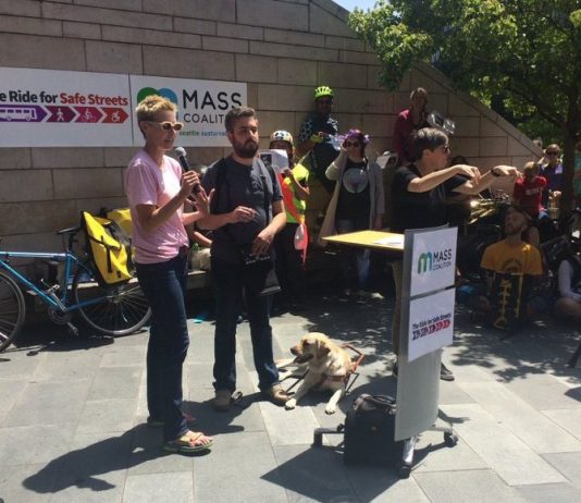 Monthly Meetup This Tuesday Featuring the MASS Coalition Anna Zivarts hold the microphone at a podium next to a bearded man with a guide dog with a crowd of people in bicycle helmets behind. Signs read "MASS Coalition" and "Ride for Safe Streets"