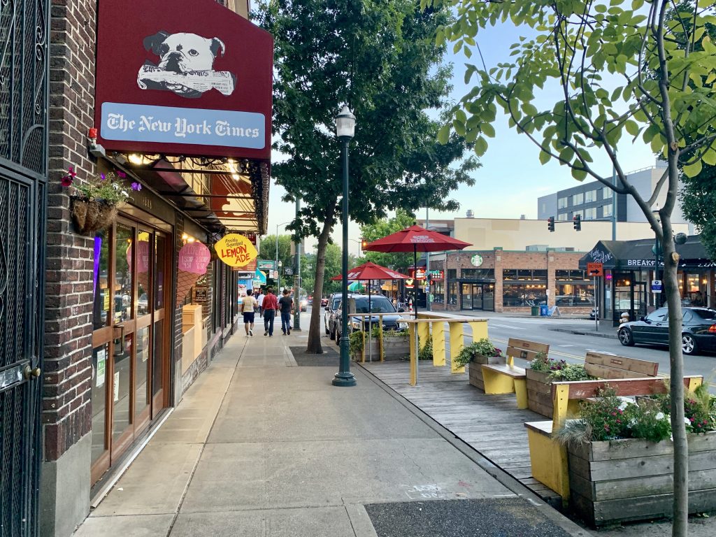 A photo showing store fronts, sidewalk, and a parklet on University Way. 