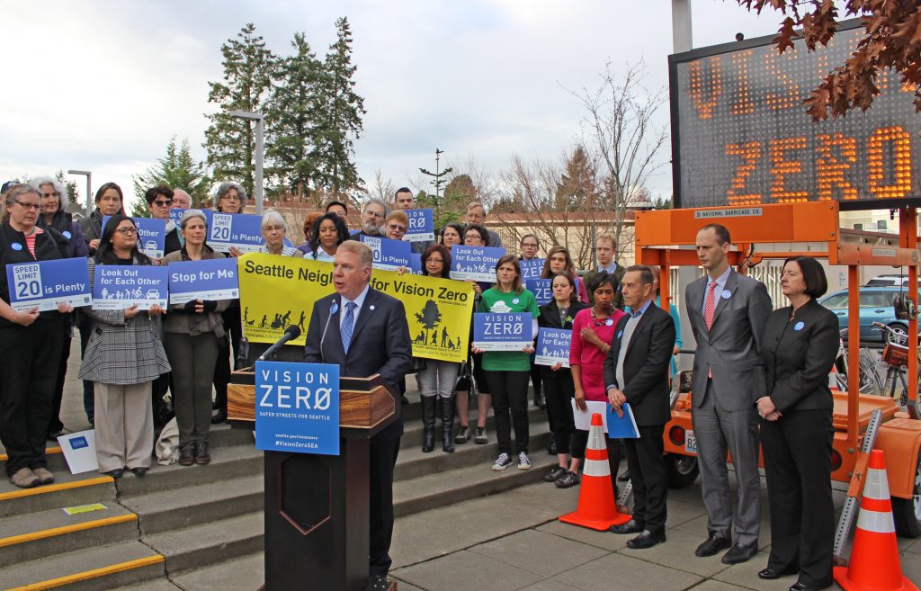 Mayor Ed Murray stands at a podium behind a crowd of staff and advocates with safety-themed signs. An electronic street signs also displays "Vison Zero."