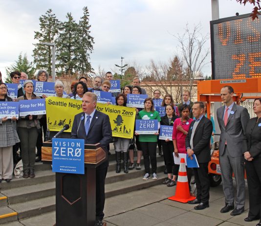 The Radical Promise of Vision Zero Mayor Ed Murray stands at a podium behind a crowd of staff and advocates with safety-themed signs. An electronic street signs also displays "Vison Zero."