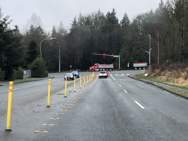 A three-way intersection of four lane roads coming to a traffic light surrounded by trees. There is too much road capacity to keep those trees.