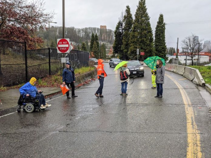 People walk and roll across the street in the rain with a car in the background.