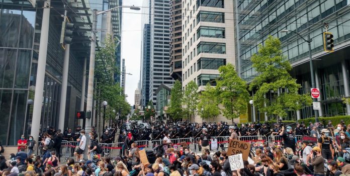 The view of Fifth Avenue with a cordon of police officers brandishing billy clubs protecting the block in front of city hall and a stream of protests headed down James Street.