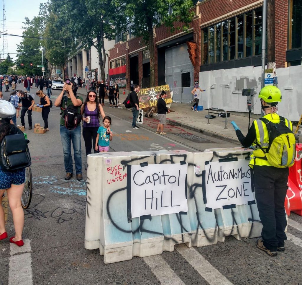 People snap pictures and wander around surrounded by street art. A construction barrier that announces "Capitol Hill Autonomous Zone" (Photo by Doug Trumm)
