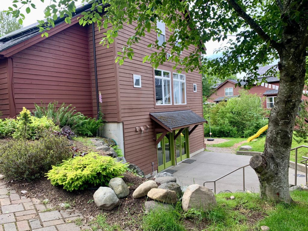 A brown wooden cohousing complex with a large tree and bushes bordering its patio.