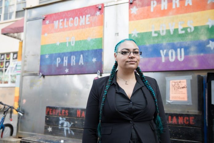 Lascelles poses in front of a rainbow flag reading "Welcome to PHRA" and "PHRA [People's Harm Reduction Alliance] Welcomes You." Lascelles wear their signature teal braids and glasses.