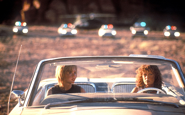 Thelm and Louise in a white convertible with cope cars in the background in the iconic scene from the 1991 film.