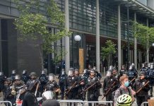 Op-Ed: Seattle Police Officers Guild is the Problem Police hold nightsticks in full riot gear behind a metal fence as protesters march by.