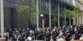 Police hold nightsticks in full riot gear behind a metal fence as protesters march by.