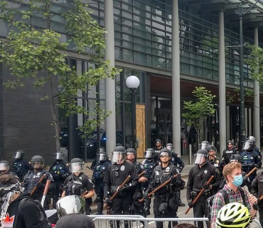 Independent Journalists Urge City to Drop Media Subpoenas Aimed at Punishing Protesters Police hold nightsticks in full riot gear behind a metal fence as protesters march by.