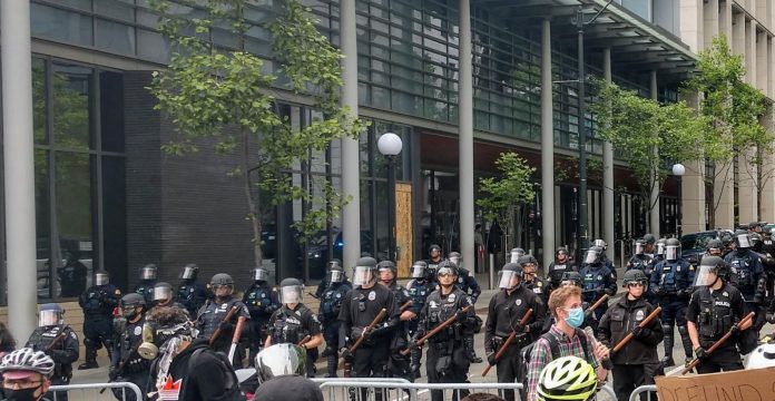 Police hold nightsticks in full riot gear behind a metal fence as protesters march by.