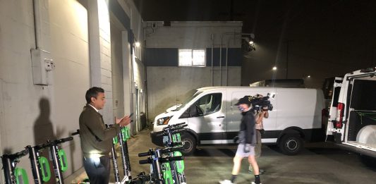 A reporter stands amidst scooters in a garage for a TV spot.