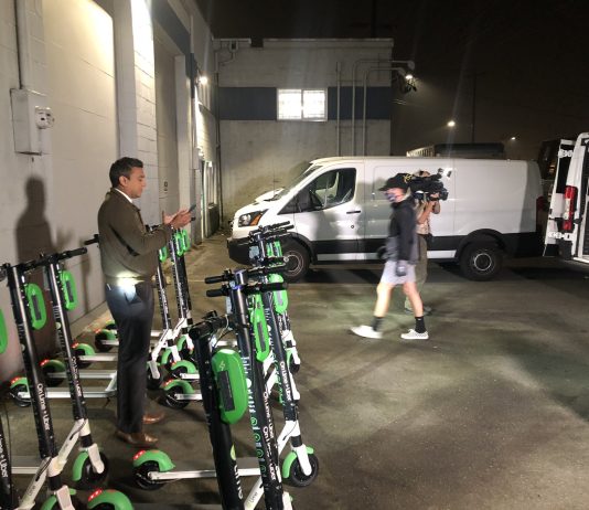Lime Rolls Out Scootershare in Seattle, LINK and Wheels a Month Away A reporter stands amidst scooters in a garage for a TV spot.