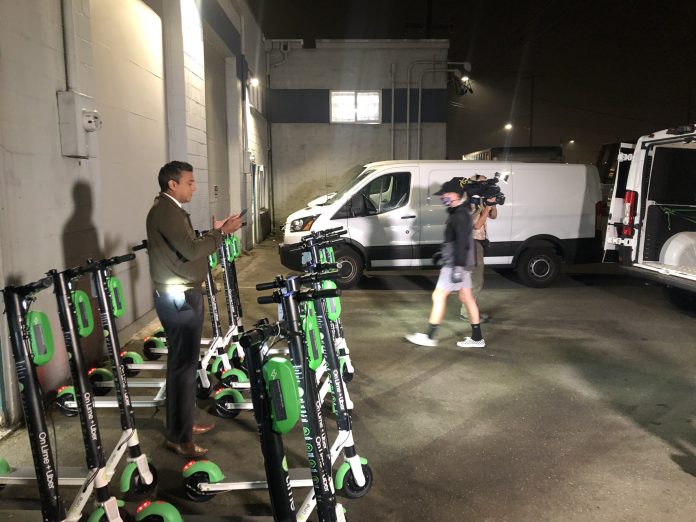 A reporter stands amidst scooters in a garage for a TV spot.