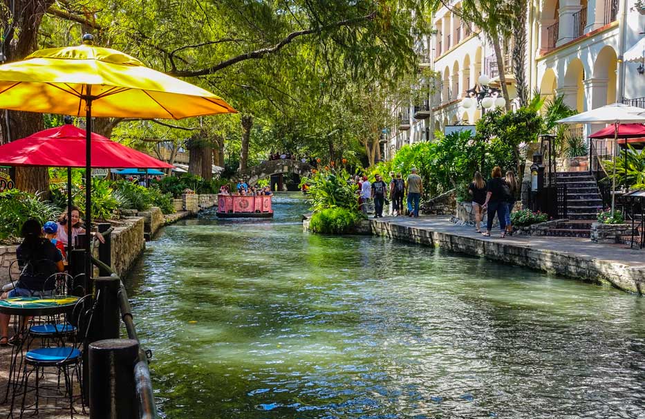 A river is flanked by umbrella and tables and trees.