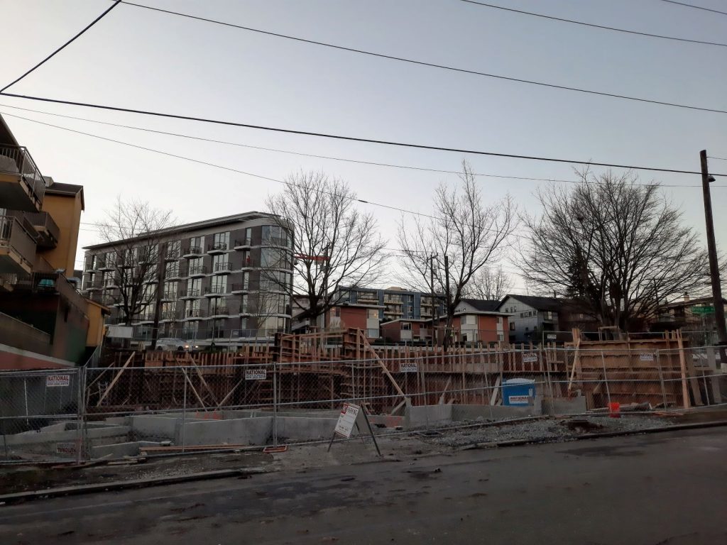 A construction pit on a triangular plot with the new Inspire Fremont building in the background across Bridge Way.